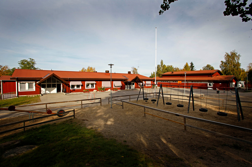 Malsta skola. Skolan har flera byggnader målade i rött med vita knutar som solen lyser på. På skolgården finns en inhägnad lekplats med sandlådor och gungor.