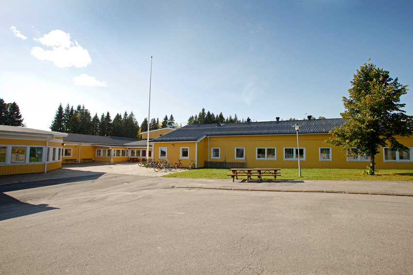 Njutångers skola. Flera avlånga gulmålade byggnader med vita knutar. Utanför skolan finns en träbänk med bord, gräsmatta med träd och en stor asfalterad plan.