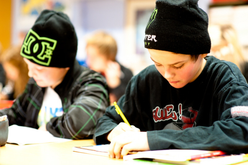 Två mellanstadiepojkar sitter och skriver i sina skolbänkar. Båda har svarta mössor med reklam på sig.