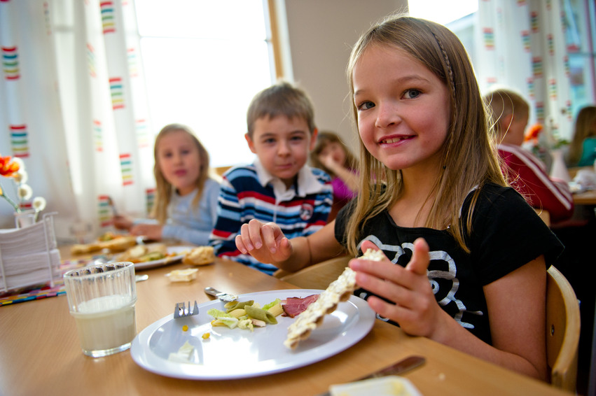 Lågstadieelever äter lunch i matsalen. En flicka håller i en hårdbrödmacka med smör på i handen och hon har nästan ätit upp maten på tallriken.