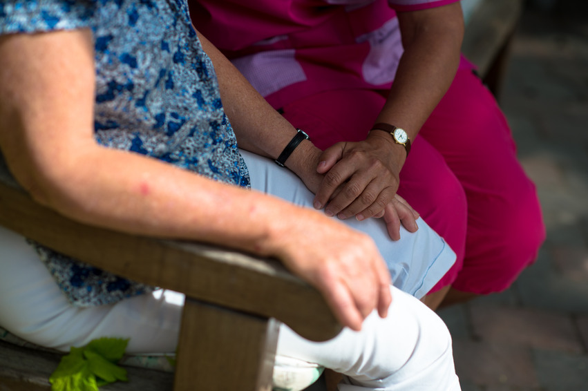 En tjej ur personalen, klädd i sina rosa arbetskläder, sitter på en uteplats och samtalar med en boende i skuggan. Hon håller sin hand ovanpå den äldre damens hand.