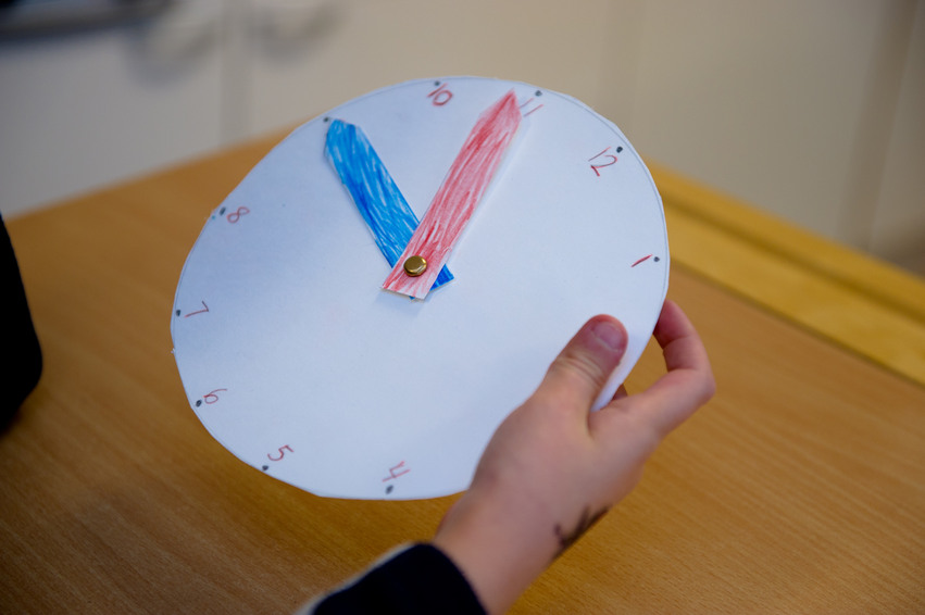 En elev sitter i sin skolbänk och tittar på en klocka gjord i papper. Klockan är vit med en blå och en röd visare och visar tiden kvart i elva.