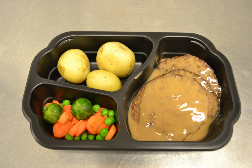 En matlåda i svart plast står på ett vitt bord.  Matlådan innehåller kokt potatis, kött med sås och grönsaker.