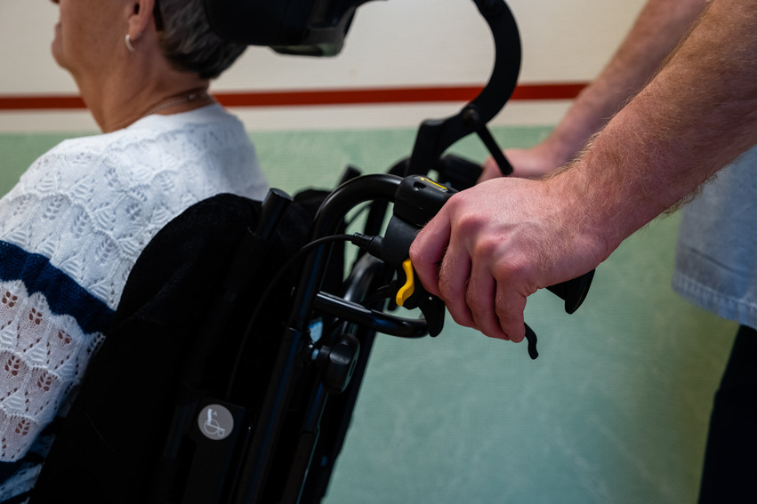En manlig anställd skjuter på en rullstol genom korridoren på ett äldreboende. I rullstolen sitter en gammal kvinna i vit kofta som bor där.