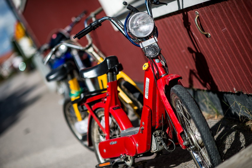 En gul cykel och en röd moped står lutad mot fritidsgården. Byggnaden är röd med vita knutar. På väggen under ett fönster sitter ett fastskruvat handtag lodrätt.