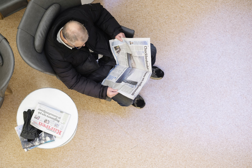 Bild tagen uppifrån på en man som sitter i grå fåtölj och läser tidningen. Bredvid står ett litet vitt runt bord och på det ligger flera tidningar.
