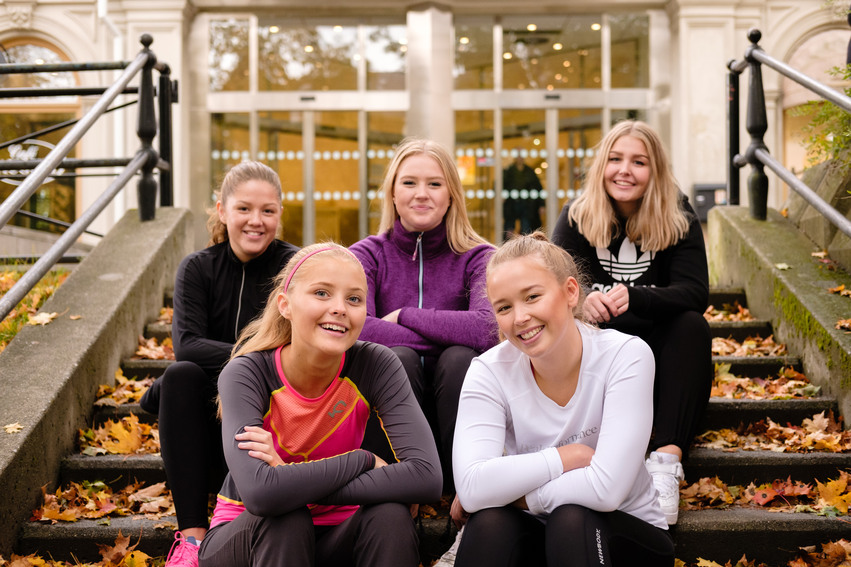 Fem yngre tjejer sitter i en trappa utomhus utanför Kulturhuset i Hudiksvall. Stentrappan är full med löv i vackra höstfärger.