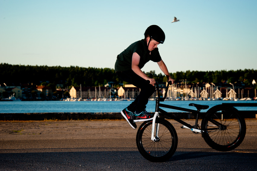 En ung kille står på framhjulet och balanserar på sin cykel nere på kajkanten. Han har en svart hjälm på huvudet.