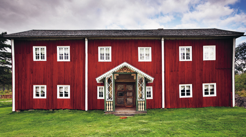 En stor Hälsingegård i Ljusdals kommun. Byggnaden är rödmålad med vita knutar och fönsterkarmar, taket är svart. Ingången ligger på mitten av husets långsida och har ett litet tak. Framför huset finns en stor grön gräsmatta. Himlen är blå men täckt av stora vita moln.
