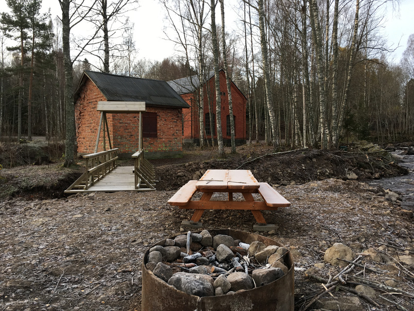 Grillplats med speciellt inrättad plats för att göra upp eld. Ett ljusbrunt träbord med bänkar på båda sidor står intill och i bakgrunden står det två små tegelbyggnader och skog. En utlagd gång i trä leder till husen.