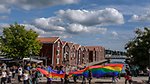 Pridetåg framför sjöbodarna i centrala Hudiksvall. Prideflaggan sveper över folksamlingen.