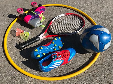 Tennisrack, fotboll, fotbollsskor, badmintonbollar och rullskridskor som ligger i en rockring på asfalt. 