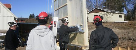 Fem personer med bygghjälmar står runt en vägg under montering.